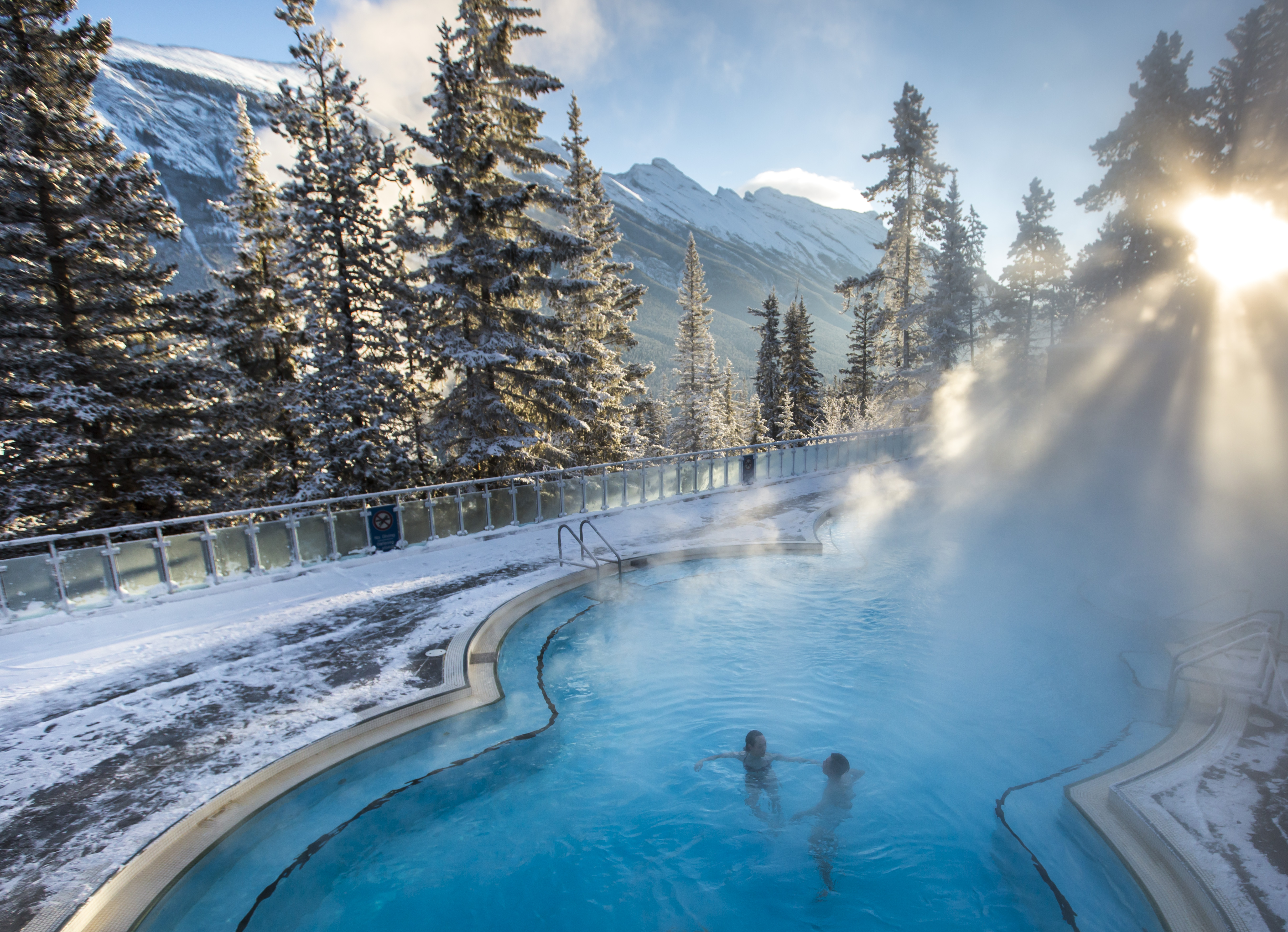 Banff Upper Hot Springs pool on a clear day with a view Mount Rundle in the distance framed by snow frosted trees