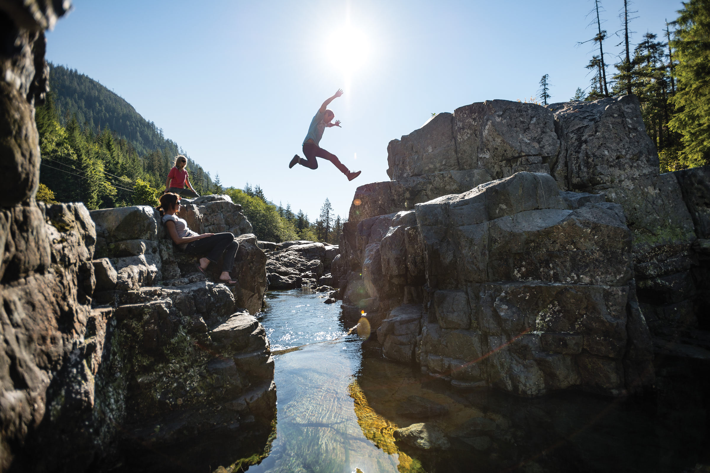 man jumps over rocky creek while friends look on