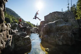 man jumps over rocky creek while friends look on