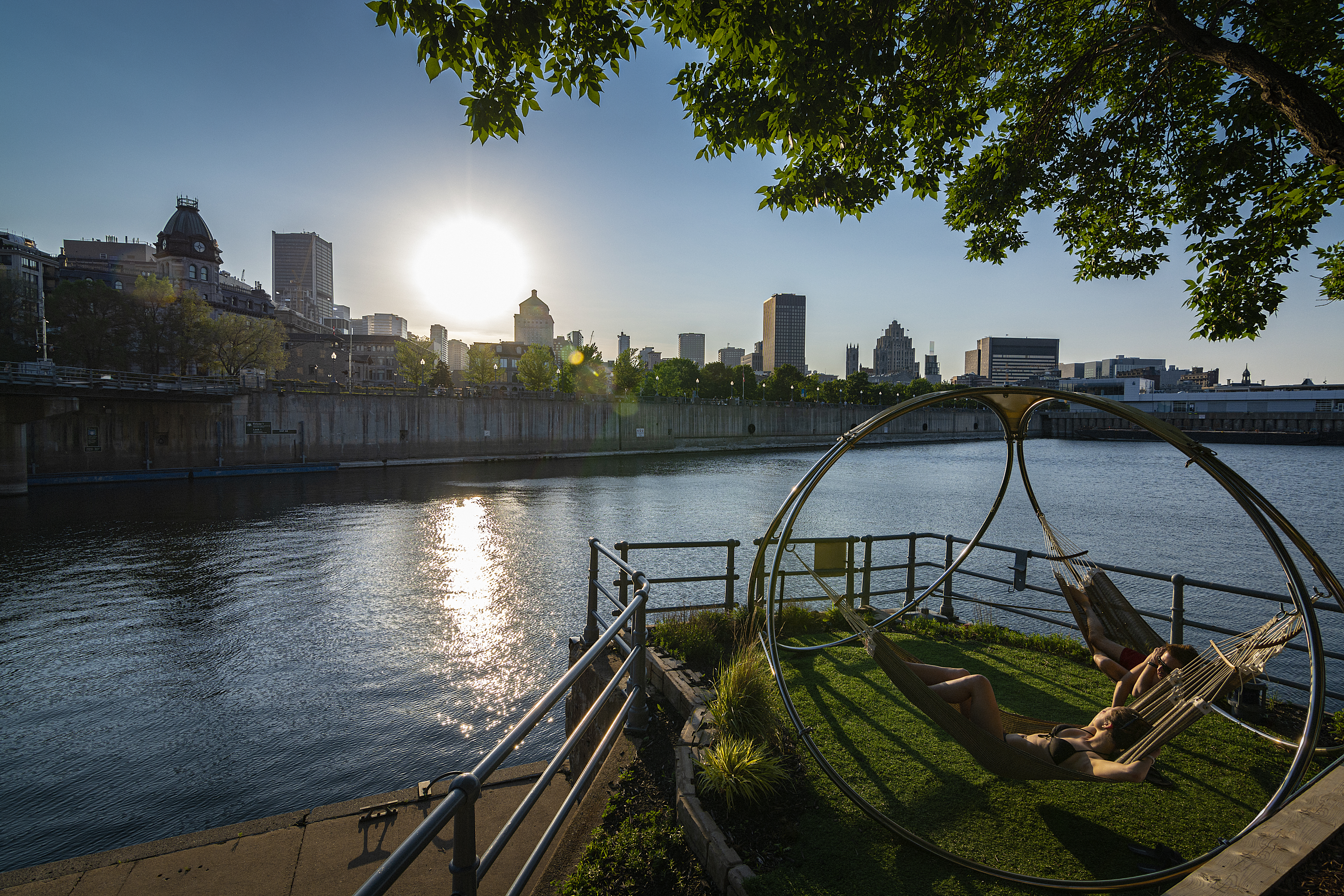 Two women sit in hammocks in an urban park with views of the St. Lawrence River and downtown Montreal