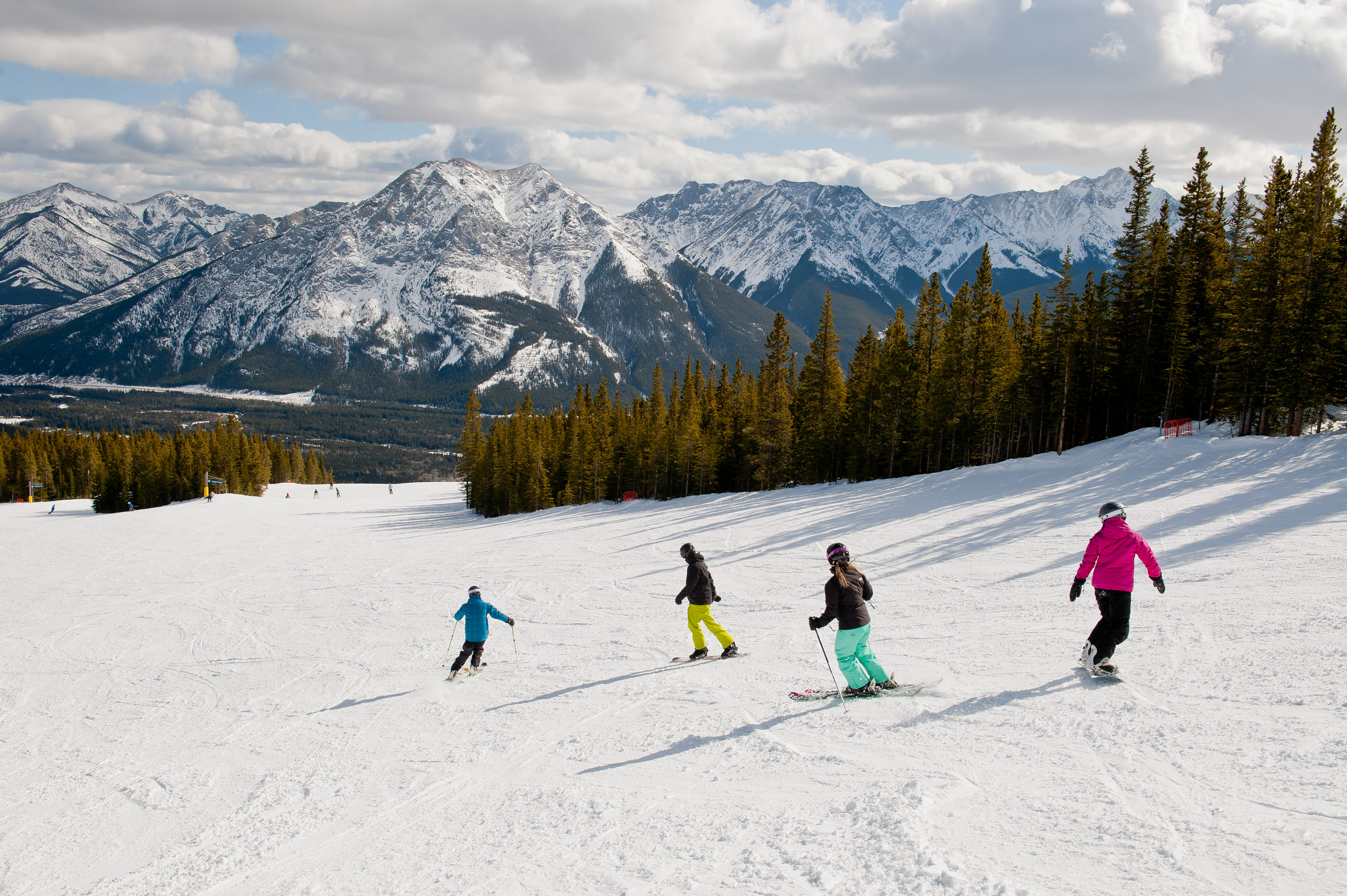 Skiers and snowboarders travel down Nakiska Ski Resort slope with open view of snowcapped mountains peaks ahead
