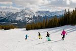 Skiers and snowboarders travel down Nakiska Ski Resort slope with open view of snowcapped mountains peaks ahead