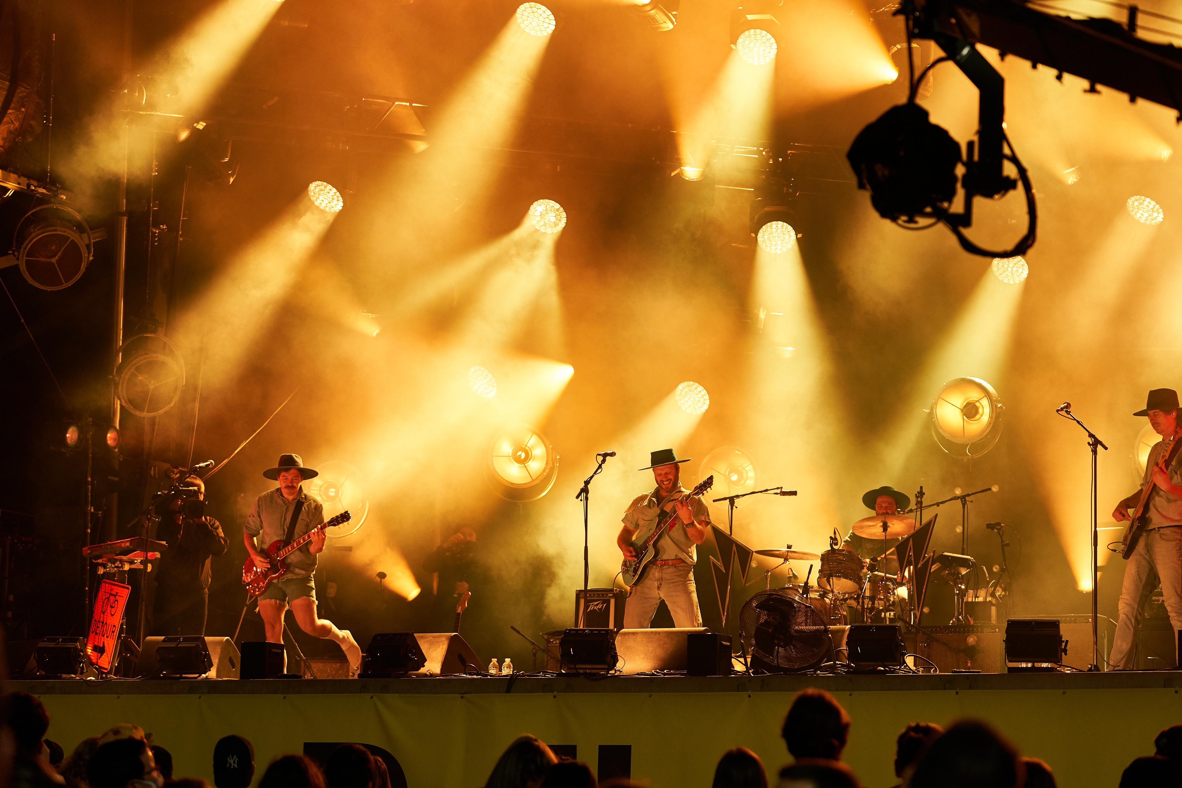 A music group, Bleu Jeans Bleu, playing guitars and drums under warm lighting on a stage