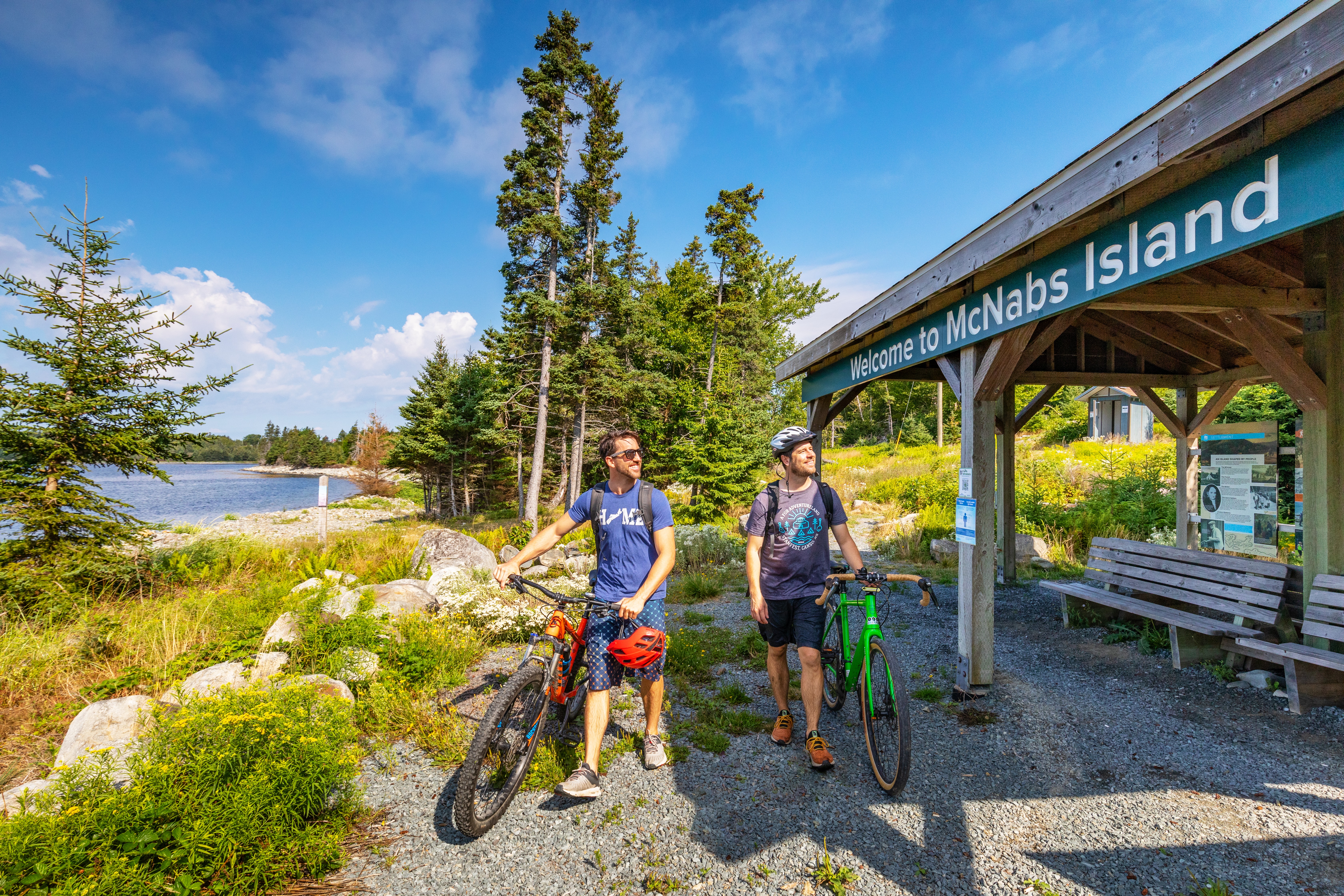 Two people walk bikes past benches and a 'Welcome to McNabs Island' sign
