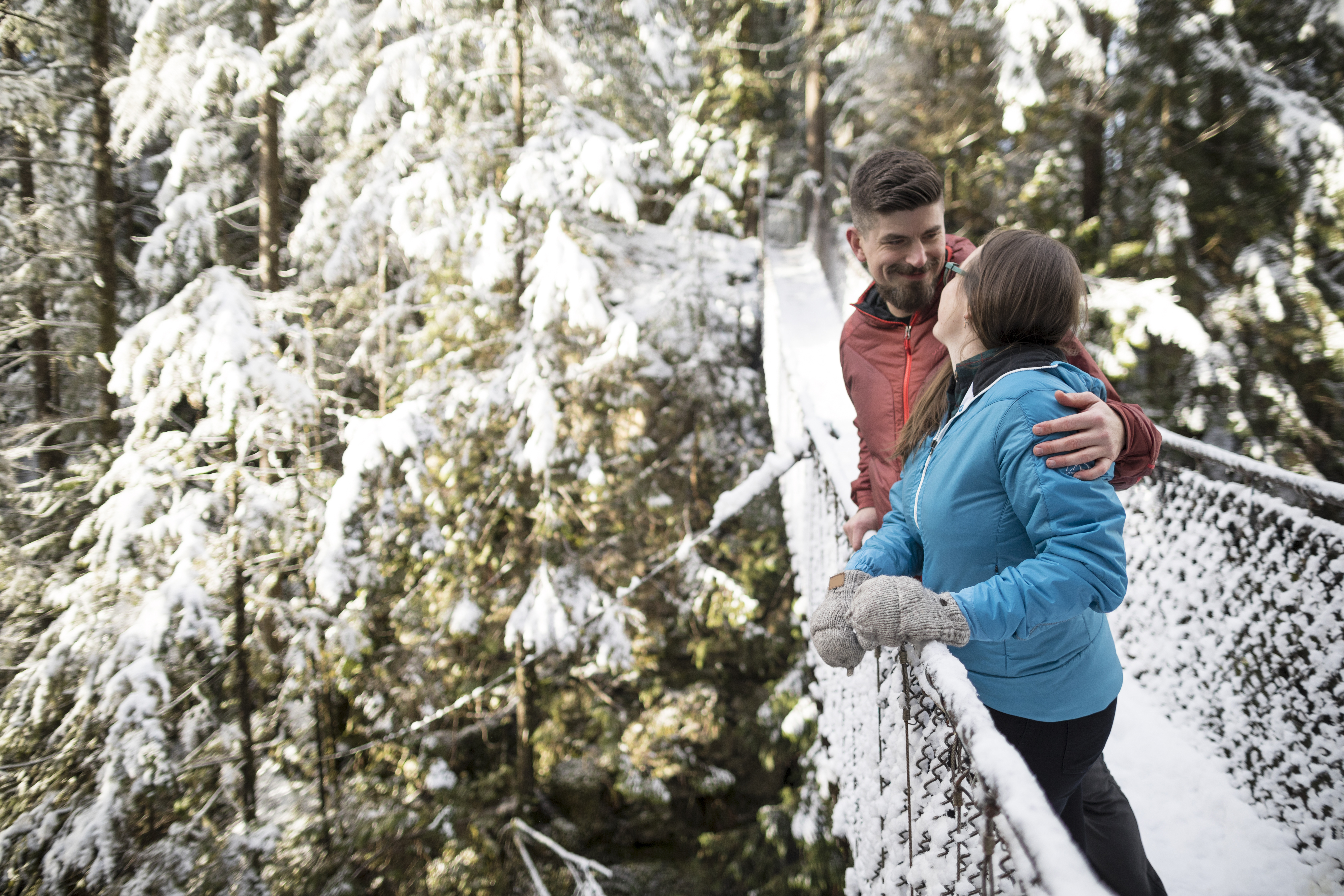 A joyful couple stand on a suspension bridge against a backdrop of tall trees covered in snow during daytime