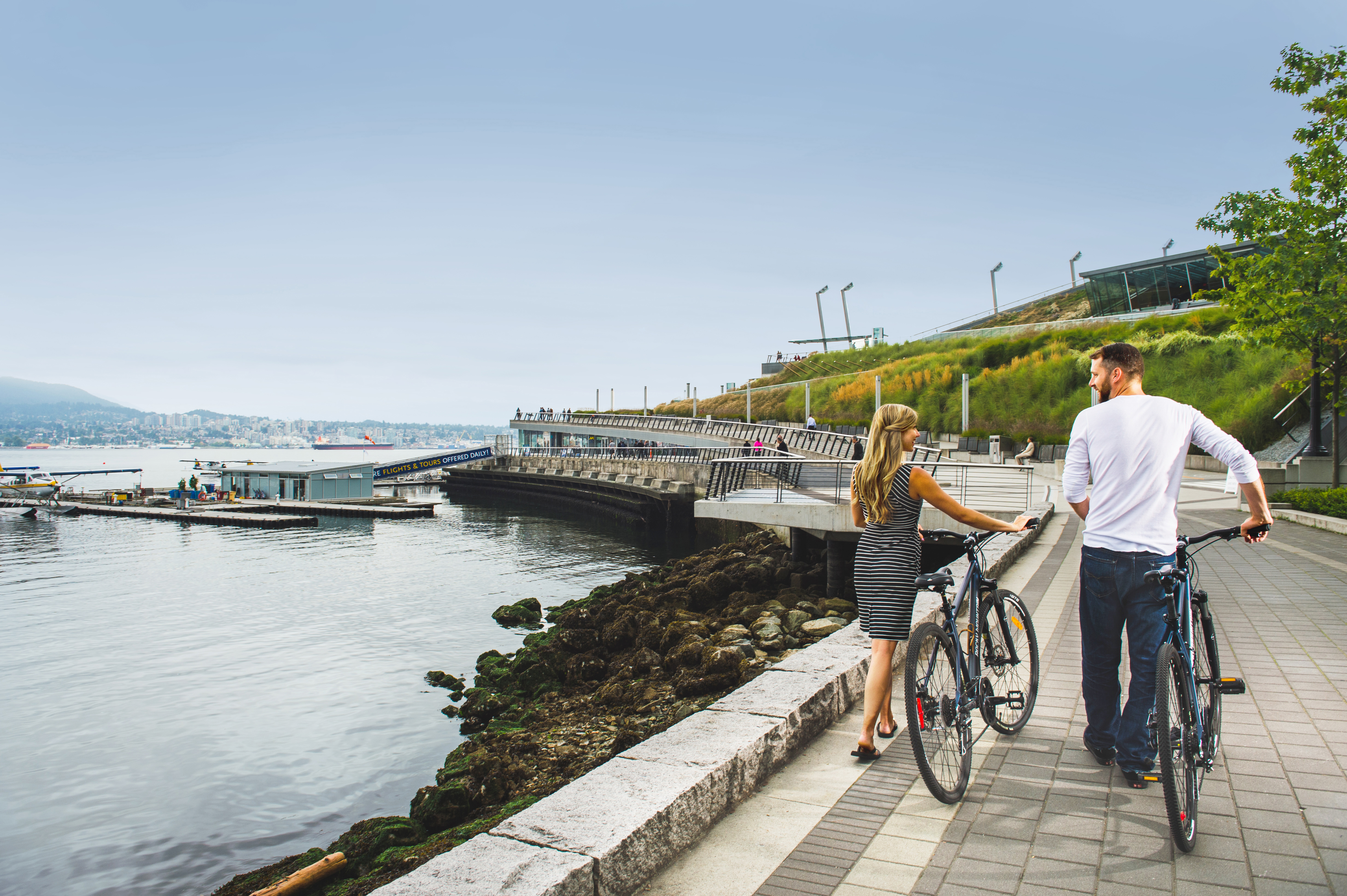 A couple walk their bikes along the sea wall in Coal Harbour with floatplanes in the background