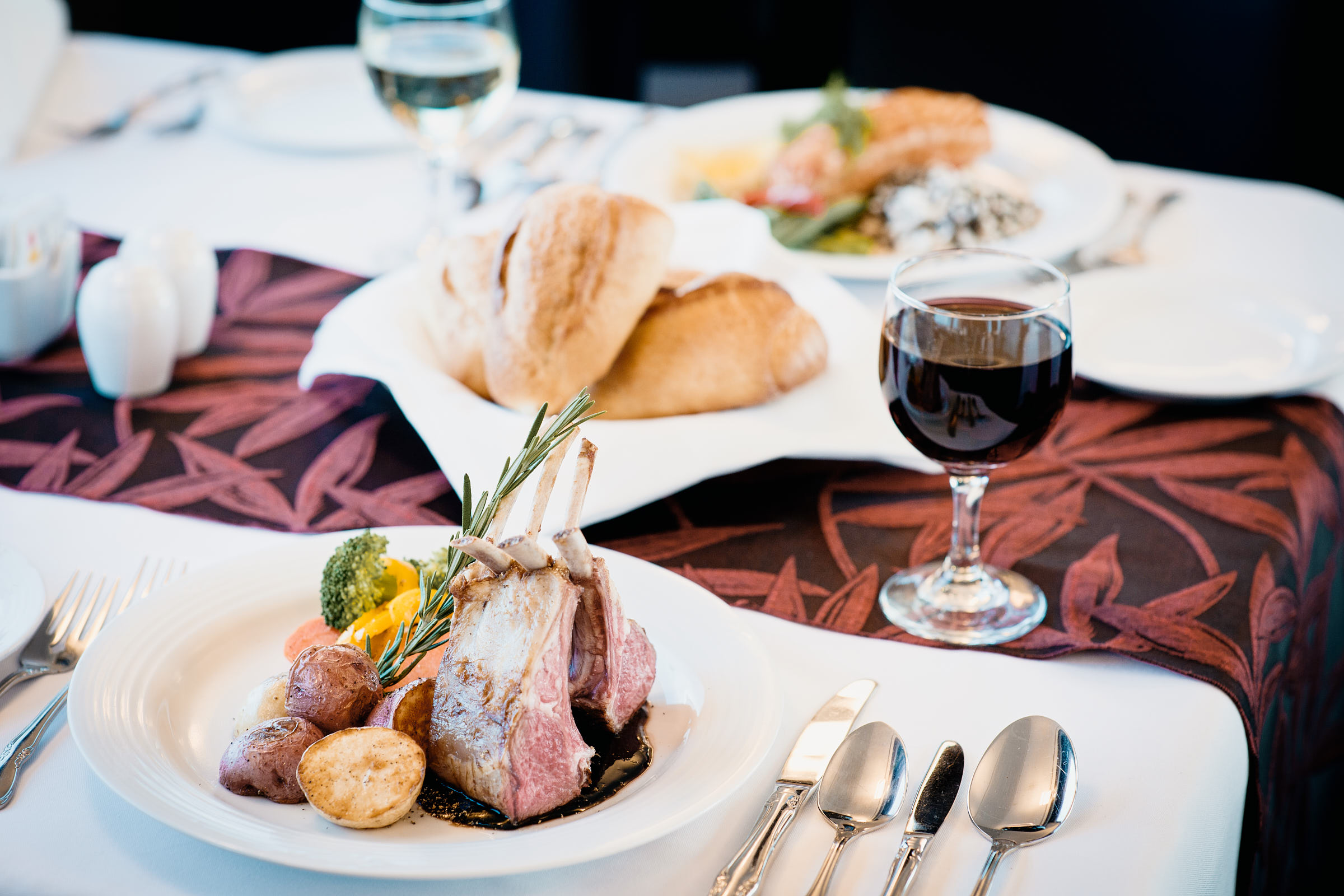 Set dinner table with a lamb rack main course, bread rolls, wine and a second main course in the background.