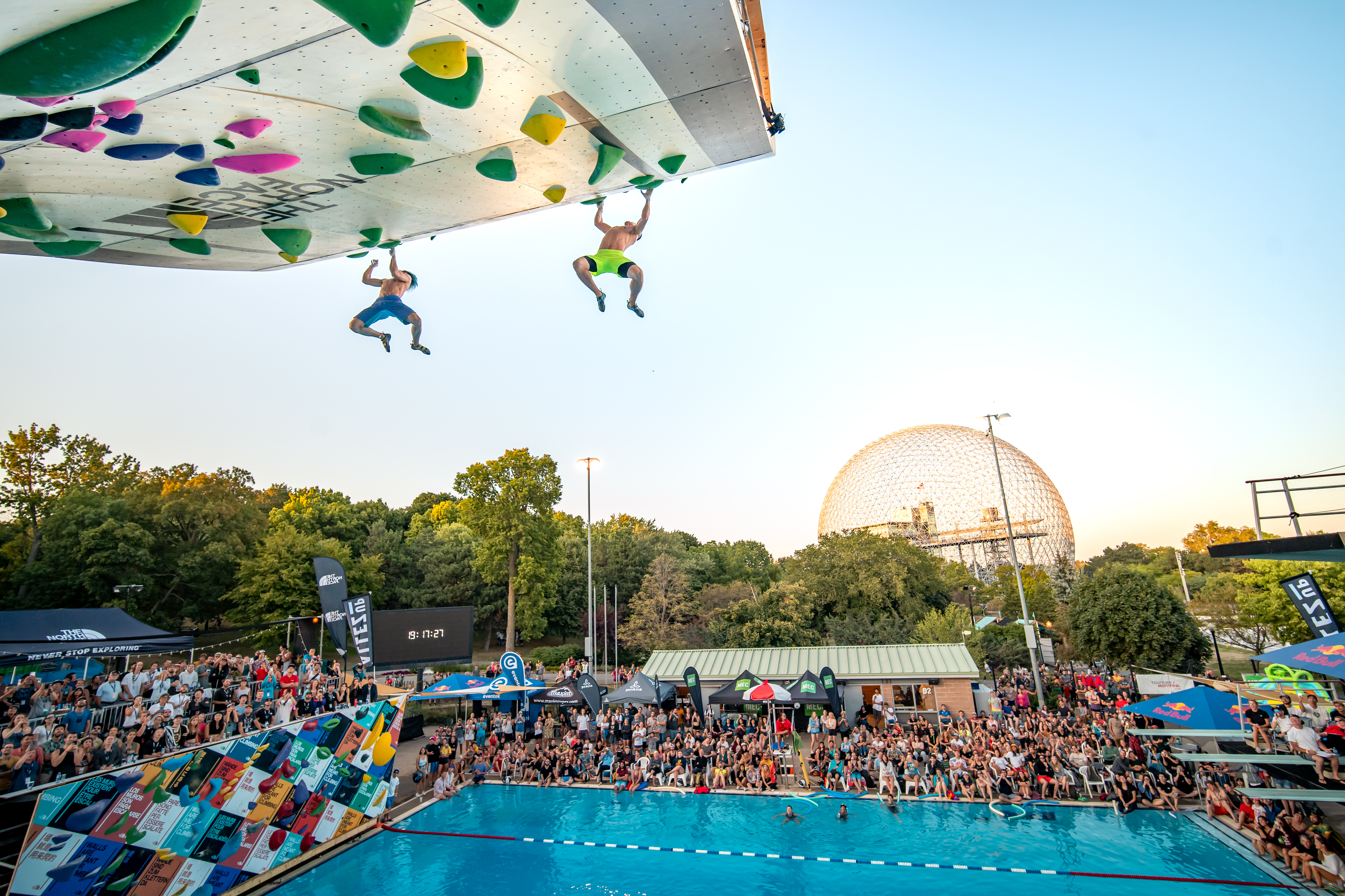 Two people free climbing on a wall overhanging a swimming pool as a crowd watches