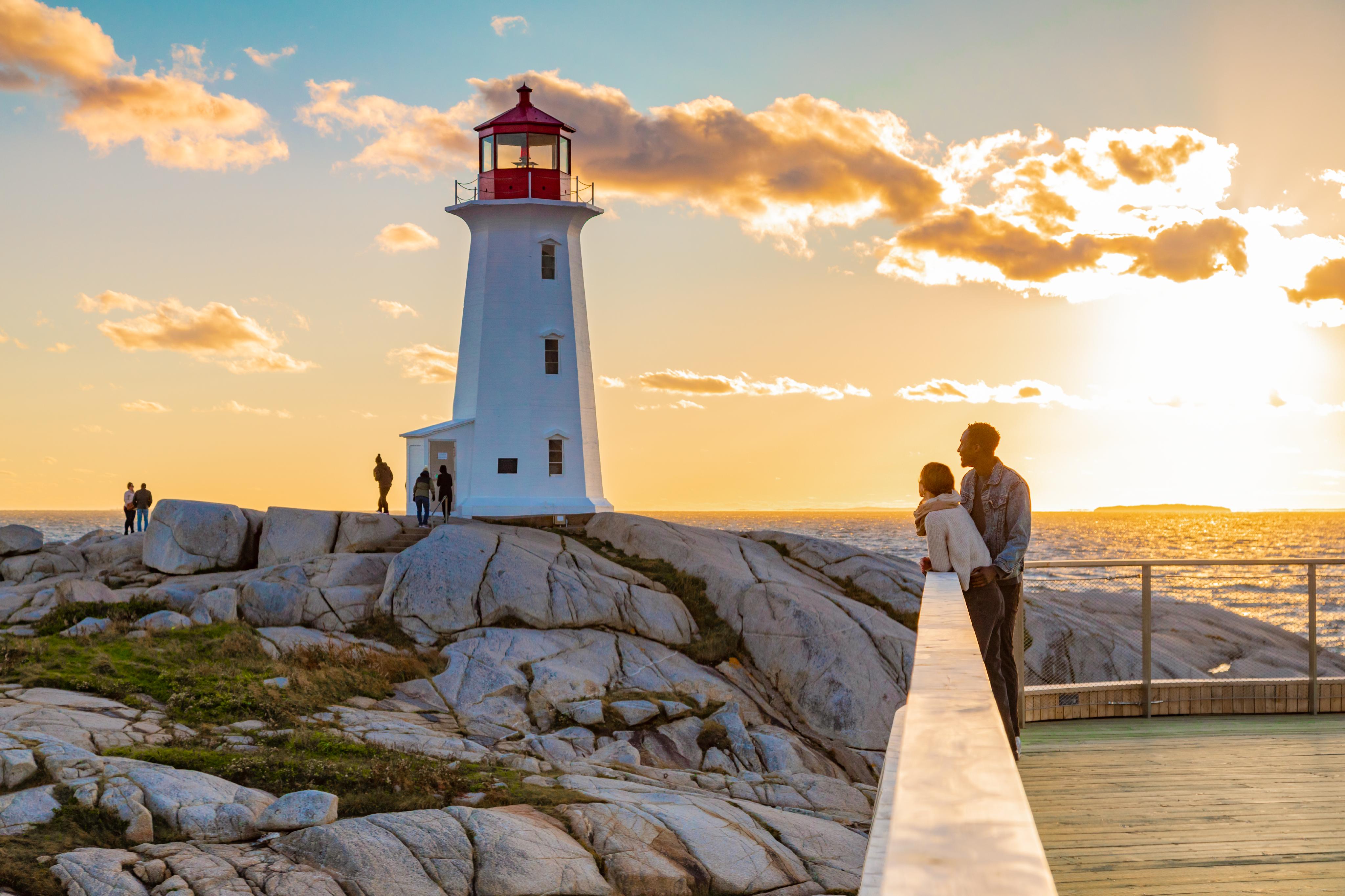 Visitors by lighthouse and shore of Peggy's Cove fishing village while the sun causes ocean water to sparkle