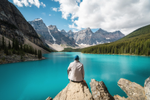 A hiker looking at unobstructed view of mountains and a dense forest while sitting on a rock above a vibrant emerald lake