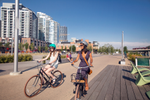 Two people riding bikes past city buildings on the Bow River pathway on a sunny day