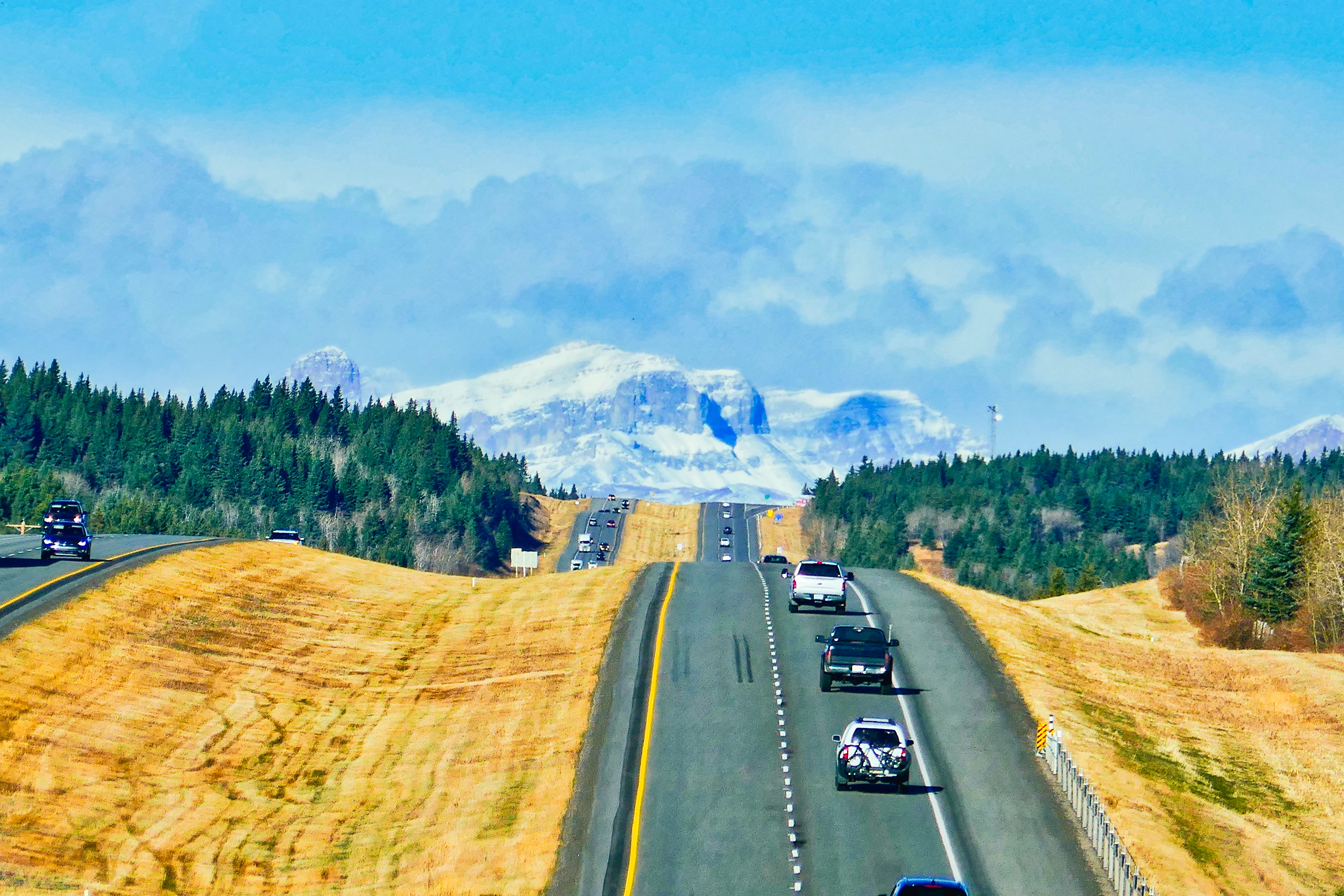 Cars on the highway with yellow grass along each side face snowcapped mountains