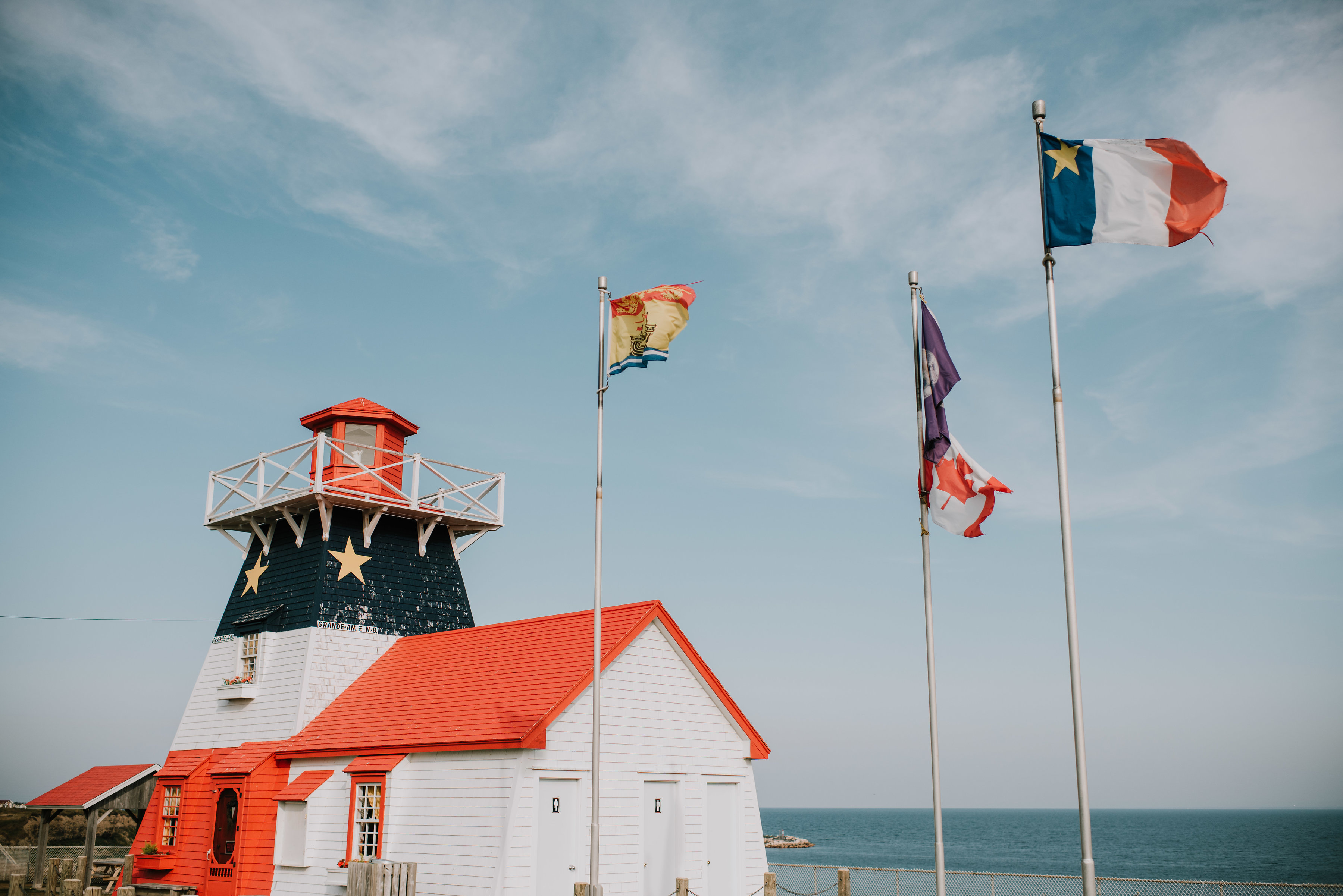 Flags fly near colourful Acadian lighthouse in coastal and seaside town, Grand-Anse, in Chaleur Bay as waves sweep