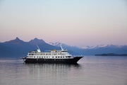 Small expedition ship sails the waters of Southeast Alaska at dusk with snowy mountains in the background.