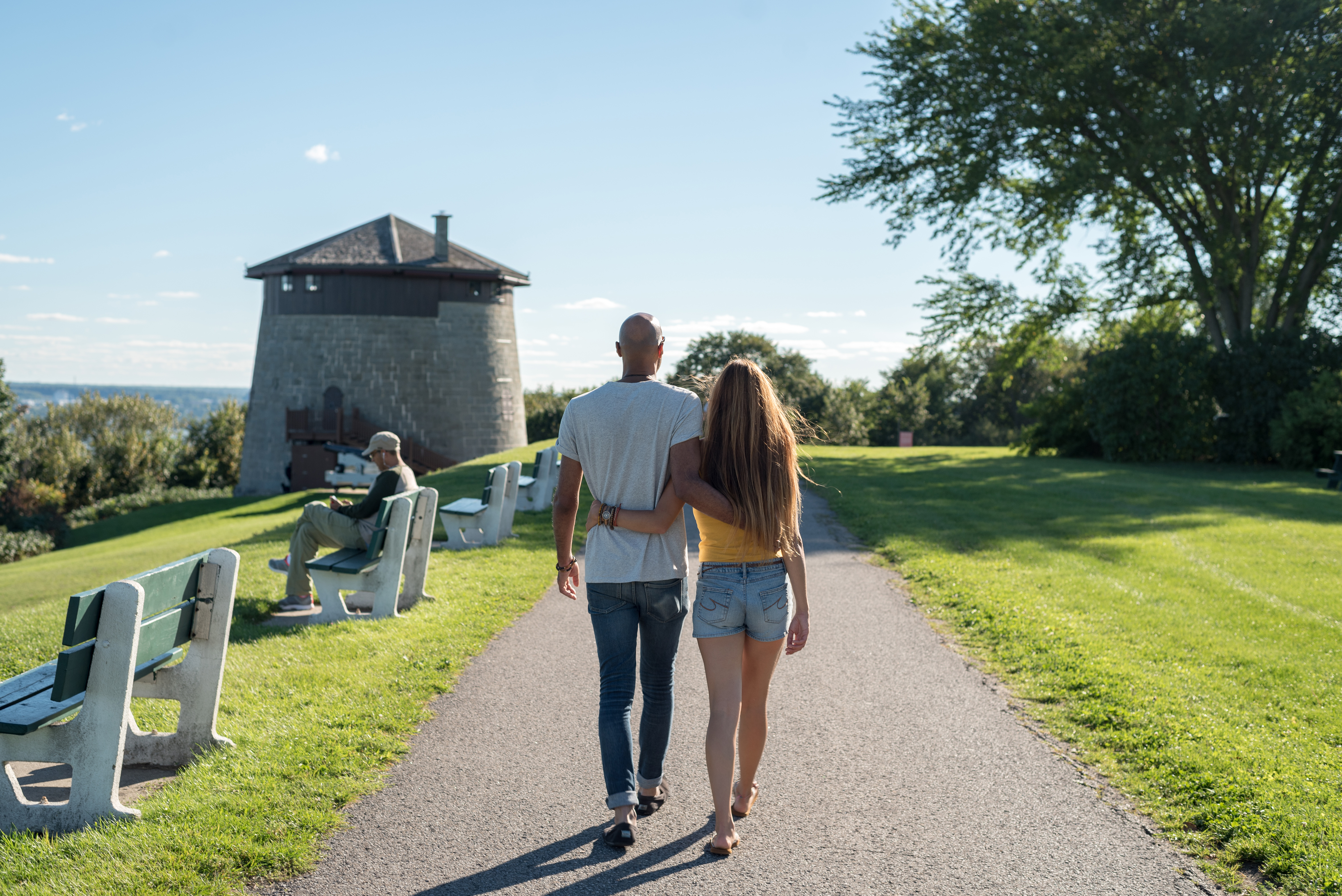 A couple walking through a park and a person sitting on a bench