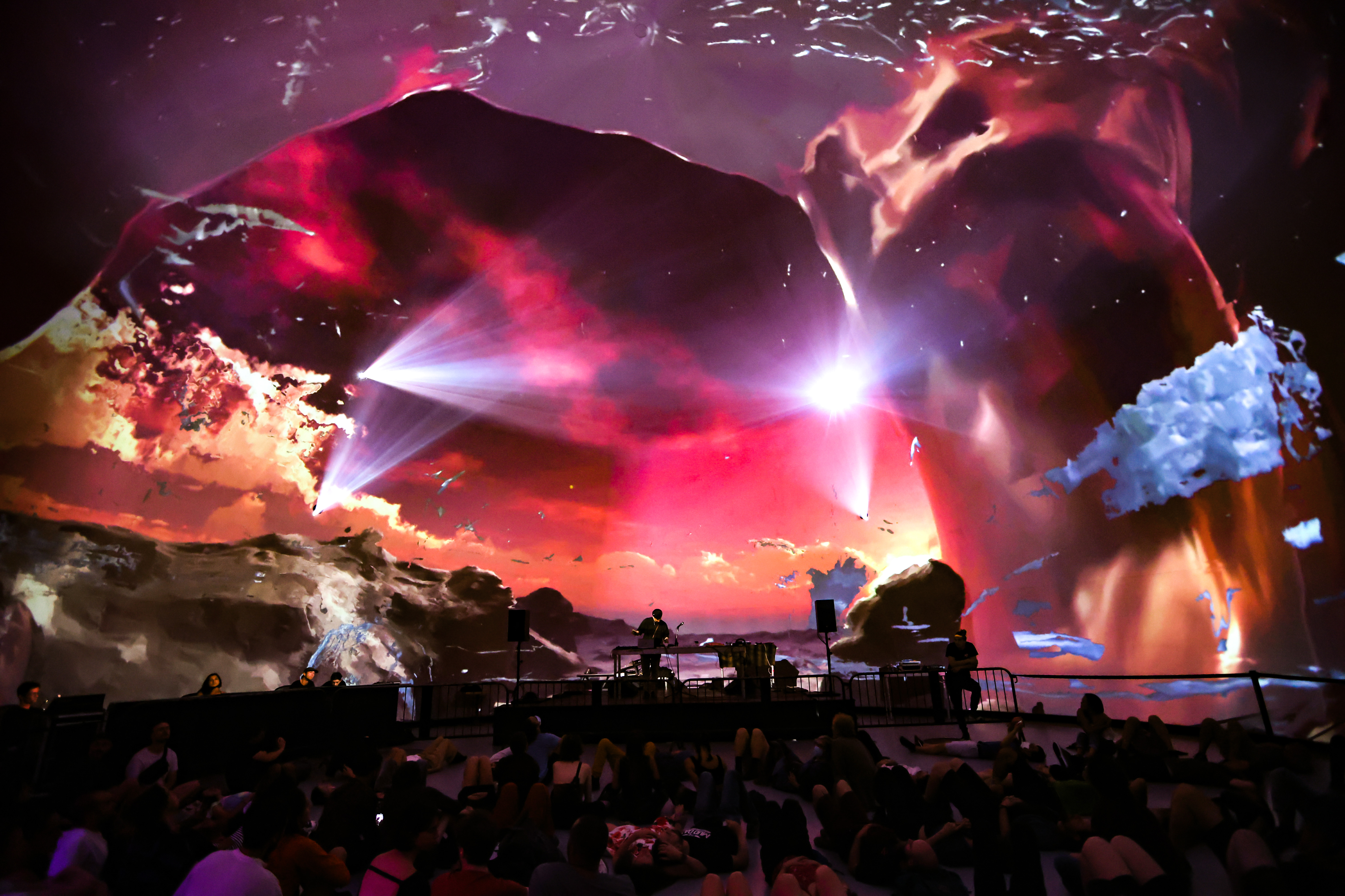 A crowd watches a musician on stage and there is a colorful digital art projection of clouds