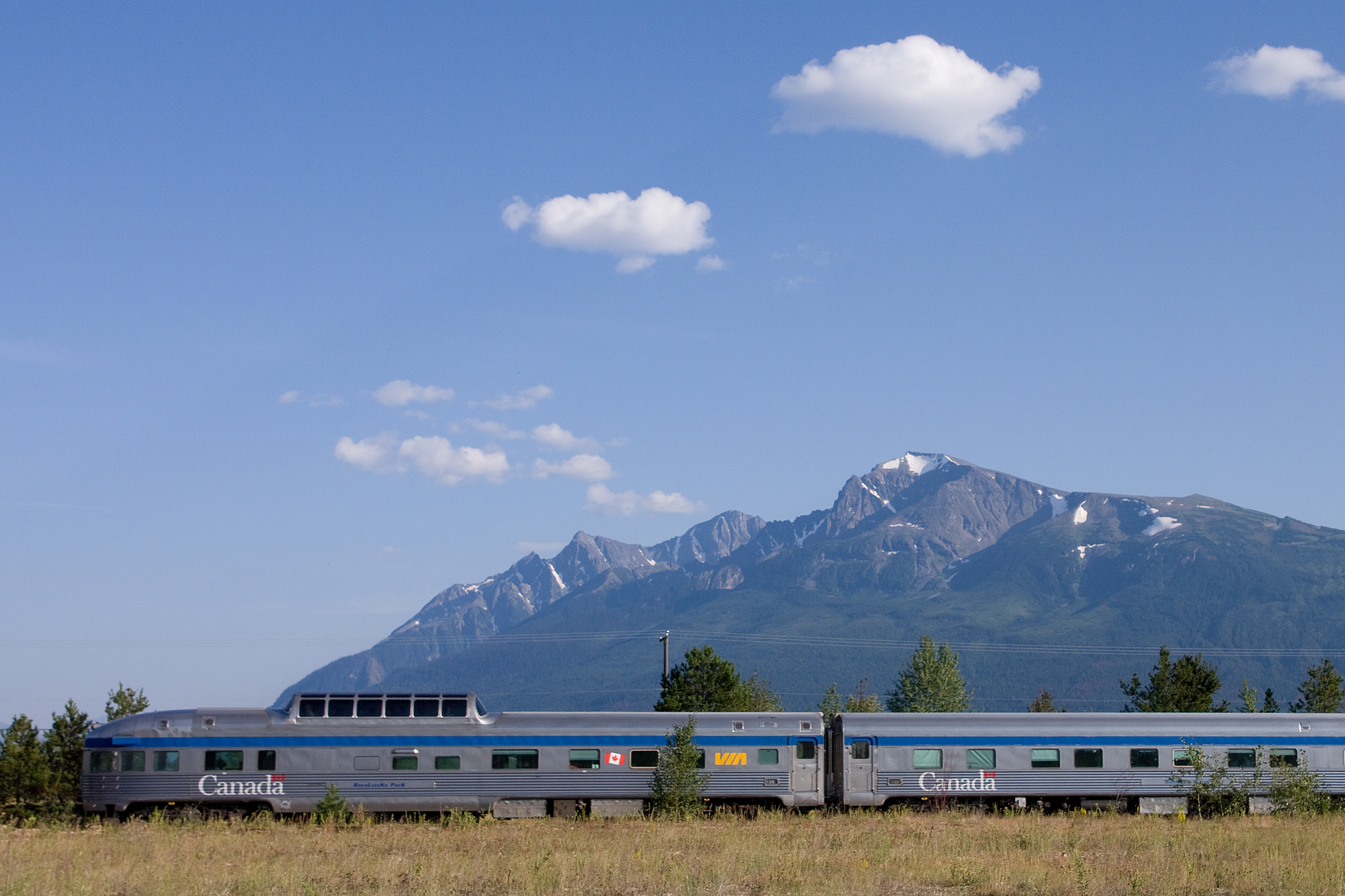 VIA Rail Canadian train travelling past mountains and trees, with grassy field in the foreground