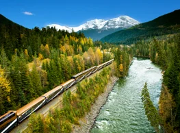 The Rocky Mountaineer train travels into the Rocky Mountains alongside a river, surrounded by fall colours.