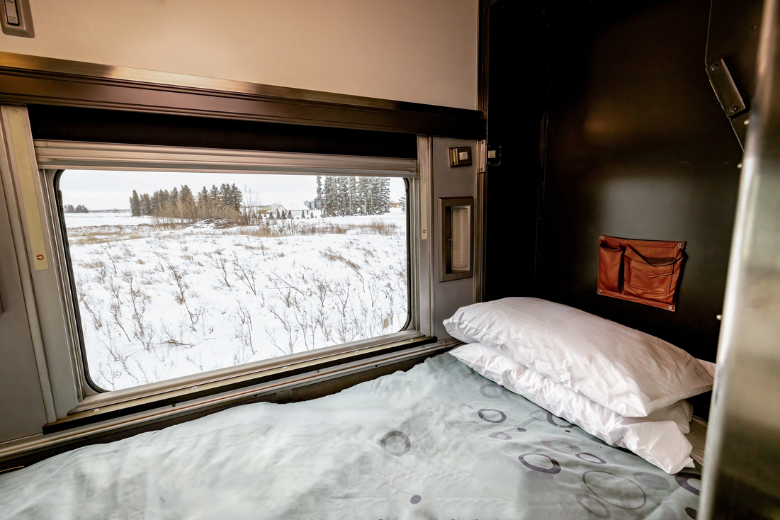 Bed and large window with blind in Sleeper Plus class cabin for one onboard VIA Rail's train, The Canadian, as it travels past snowy landscape during winter