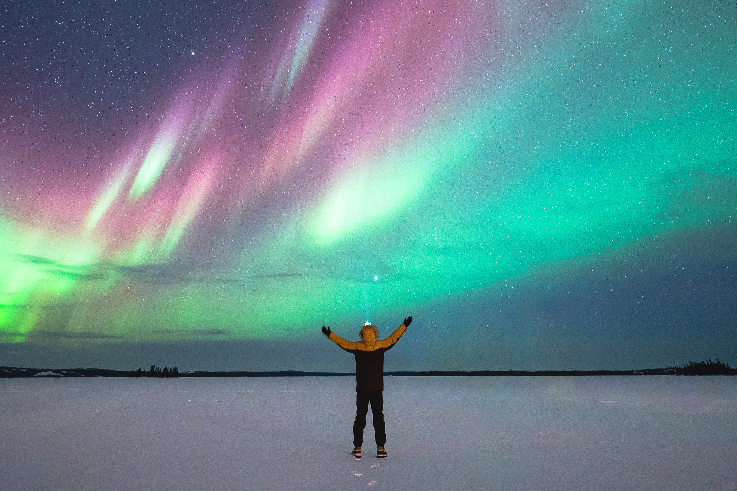 A person is standing in the snow with their arms outstretched in front of the aurora borealis