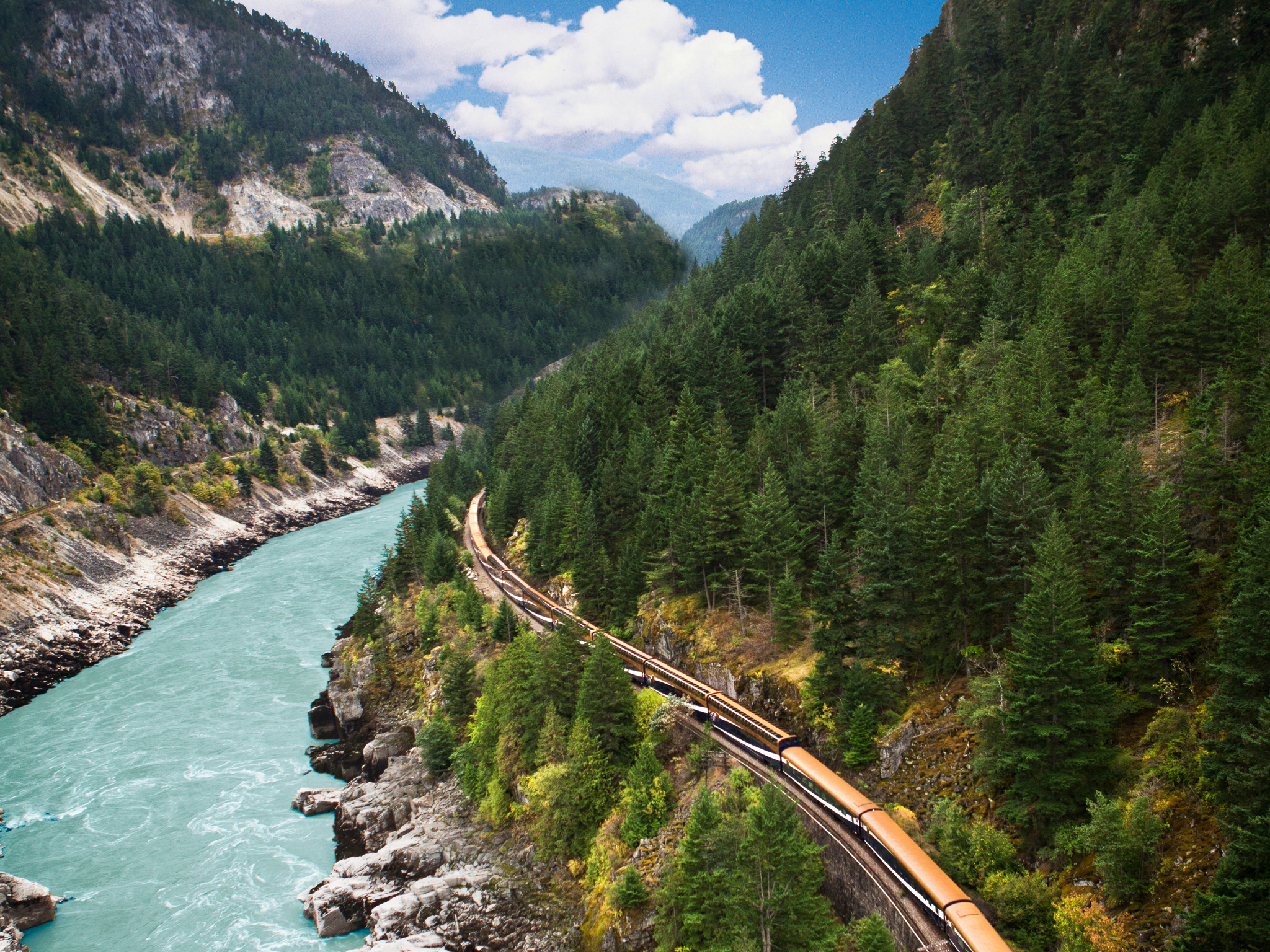 The Rocky Mountaineer train passes by a green river and lush forests.