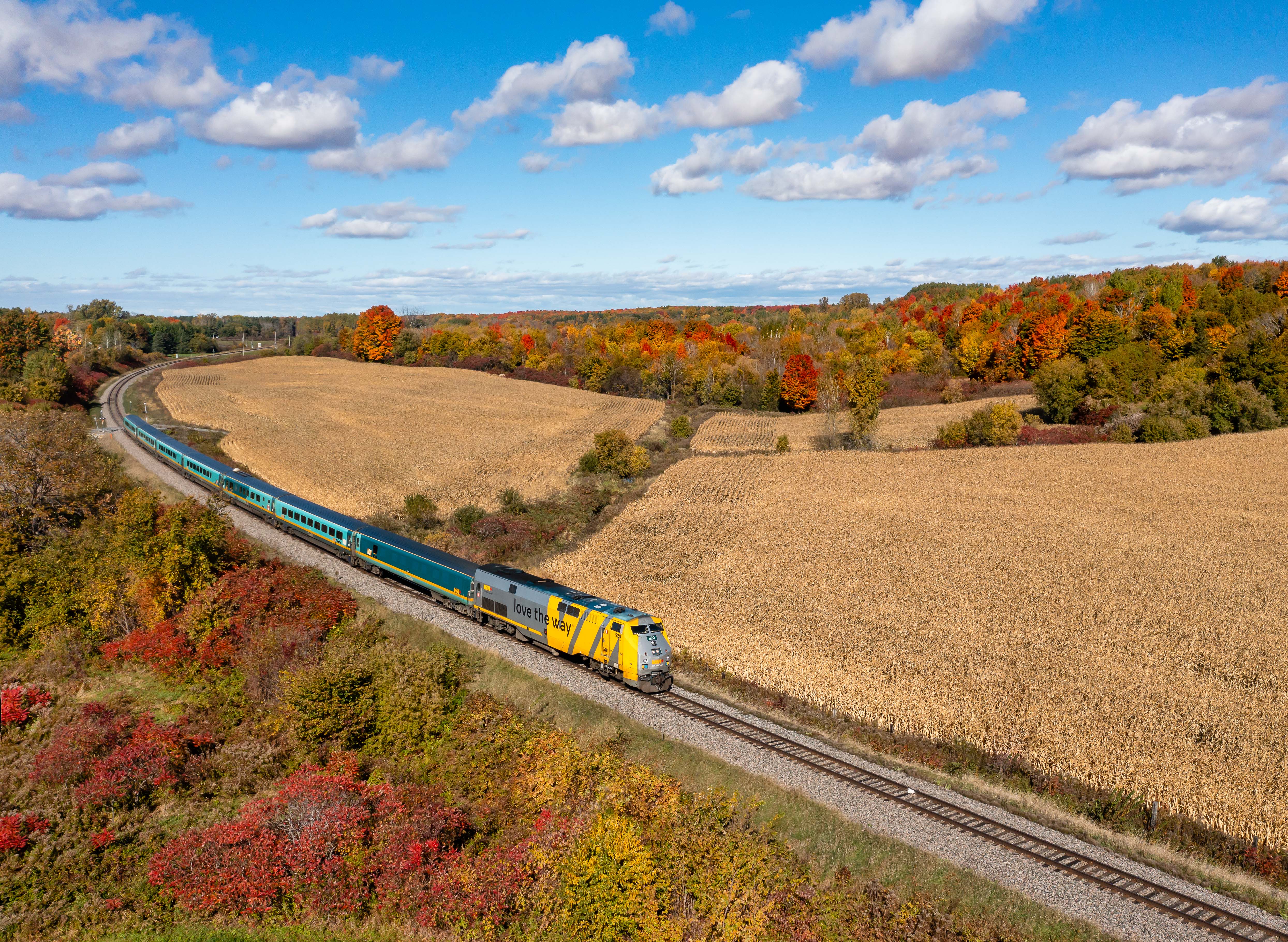 Aerial view of VIA Rail Corridor train passing fields and red fall foliage in rural Ontario