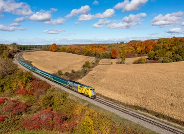 Aerial view of VIA Rail Corridor train passing fields and red fall foliage in rural Ontario