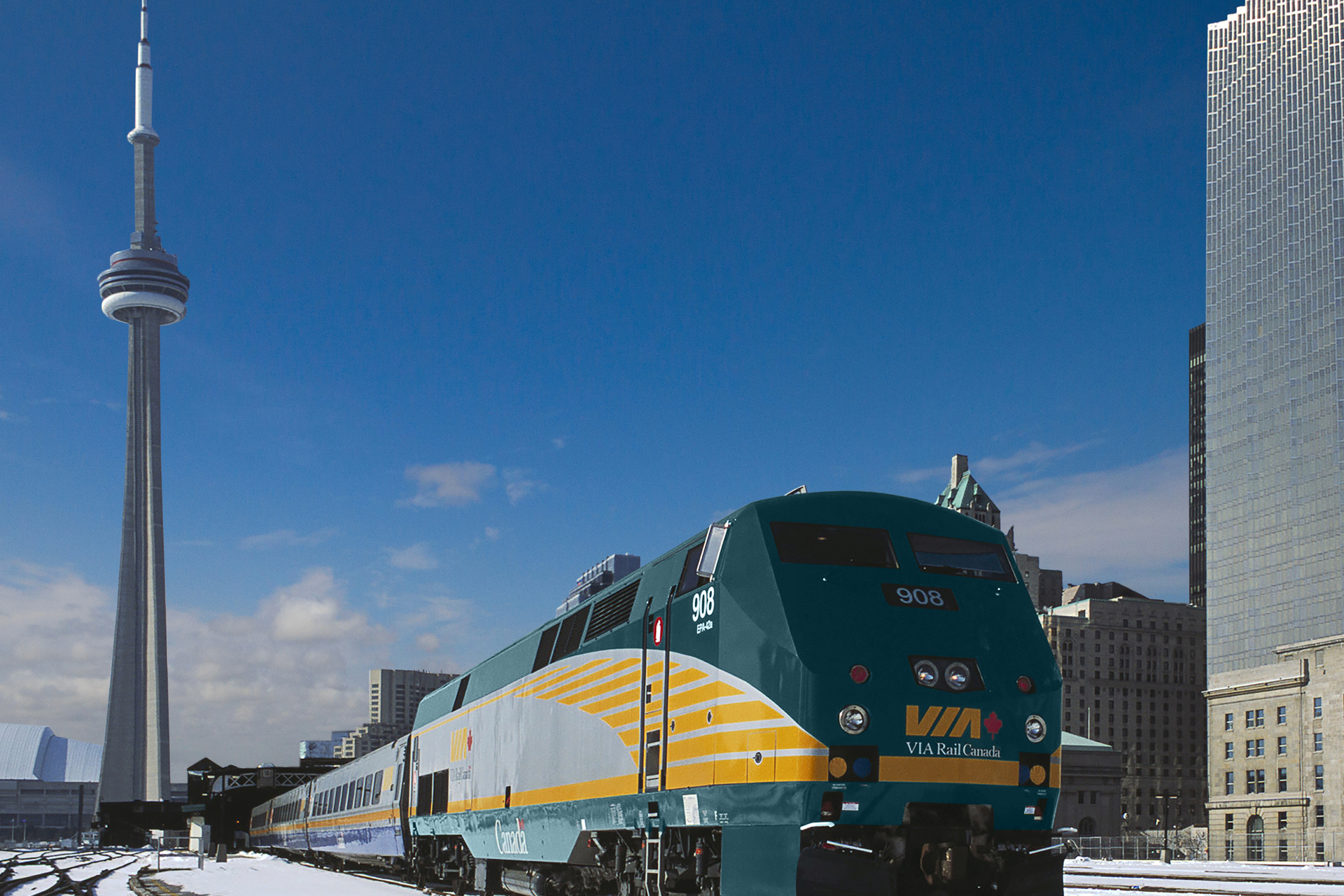 Exterior view of VIA Rail train next to a building and the CN tower in Toronto, with snow on the ground