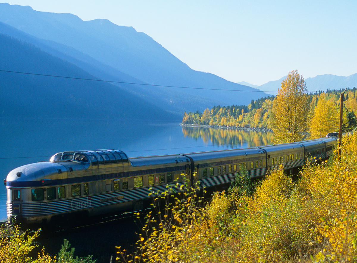 VIA Rail train travels along scenic view with fall foliage and lake with Canadian Rockies in the background