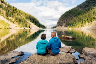 A couple sitting on a rock on the shore of Lake Agnes with mountains rising up on both sides