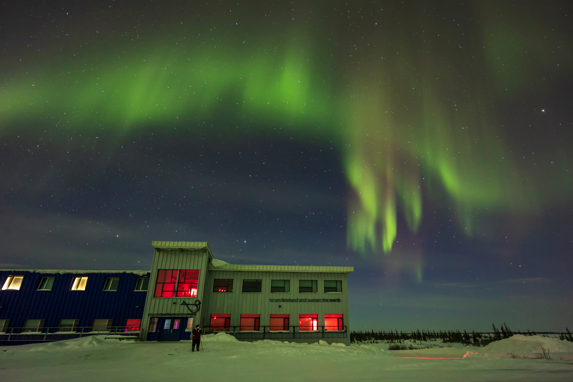 Auroral curtains in the north over the Churchill Northern Studies Centre, Canada, with a lone photographer setting up