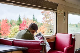 Person reading a newspaper in The Ocean train lounge, views of fall foliage from the window