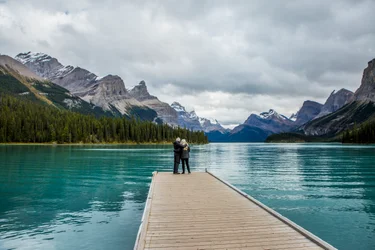mature couple standing at the end of a dock at Maligne Lake, mountains in background