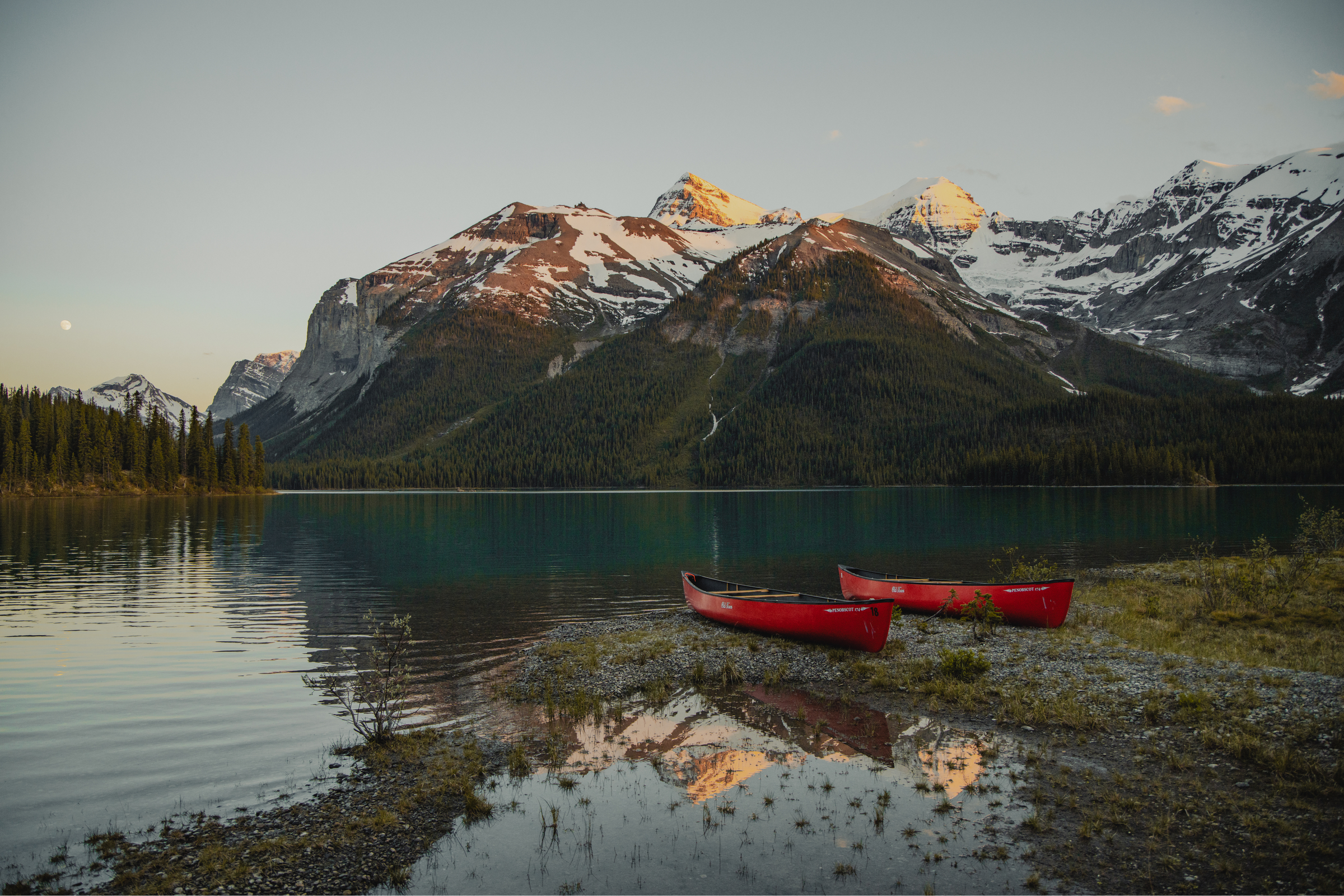 Red canoes next to a lake, with snow-capped mountains in the background, in Jasper National Park at dusk