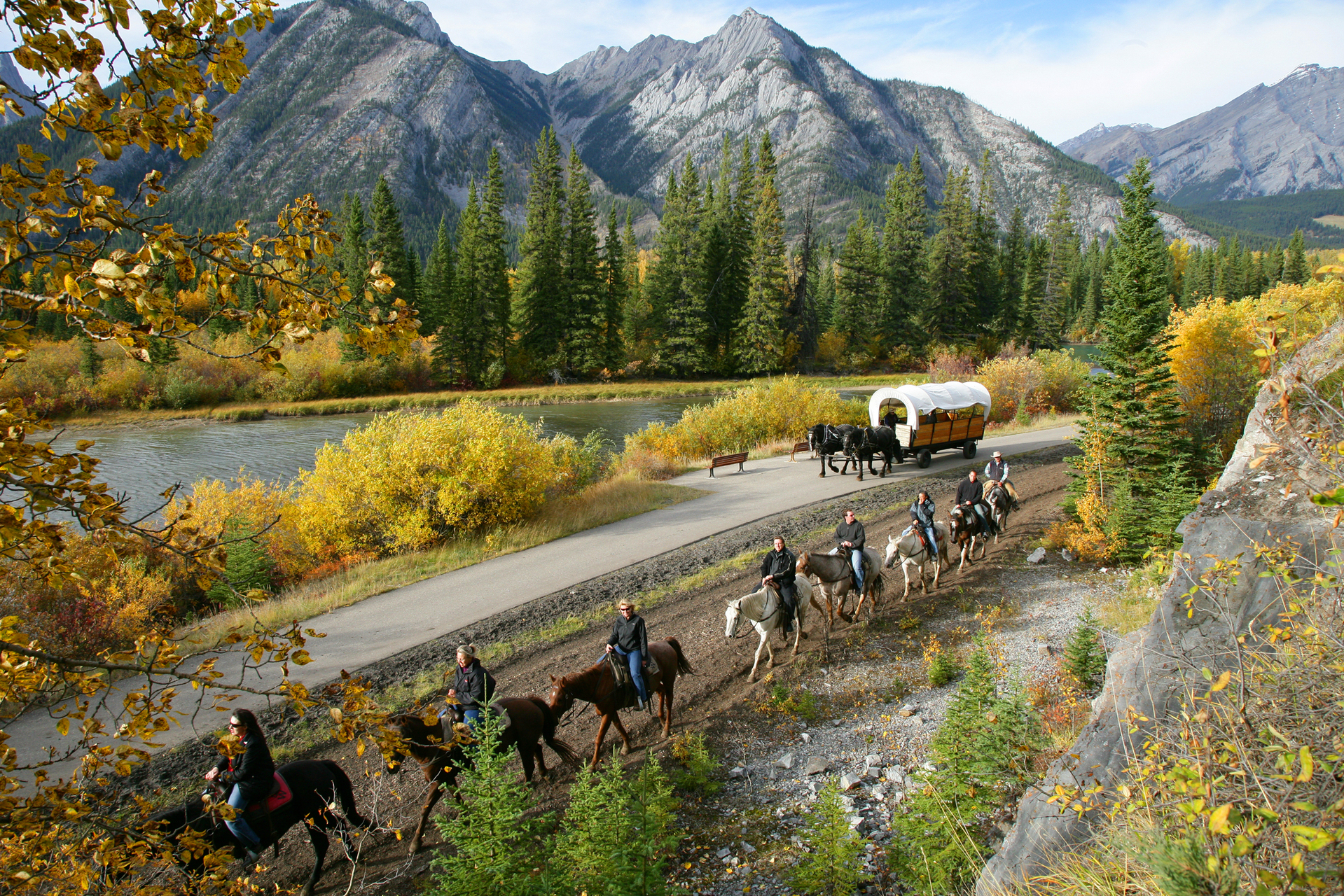 A line of people on horseback travel through the Rocky Mountains and are followed by horse and wagon 