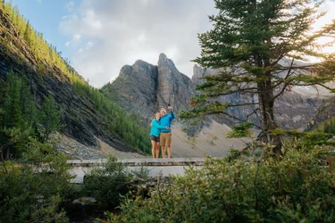 A couple take a selfie on a bridge with mountains in the background at Lake Agnes in Lake Louise