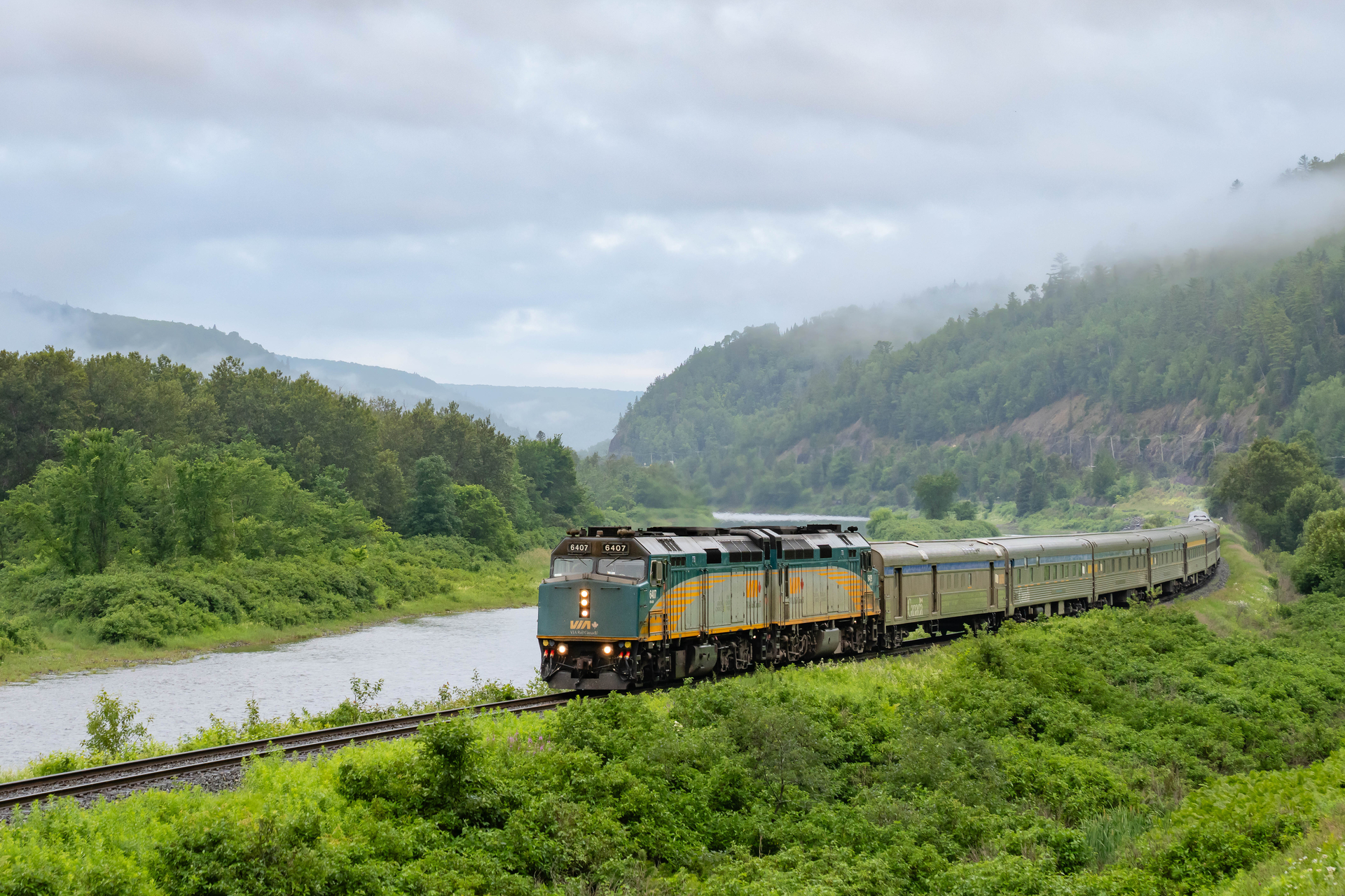 VIA Rail Ocean train rounds a corner in rural Quebec, lush green forests and cliffs in the background