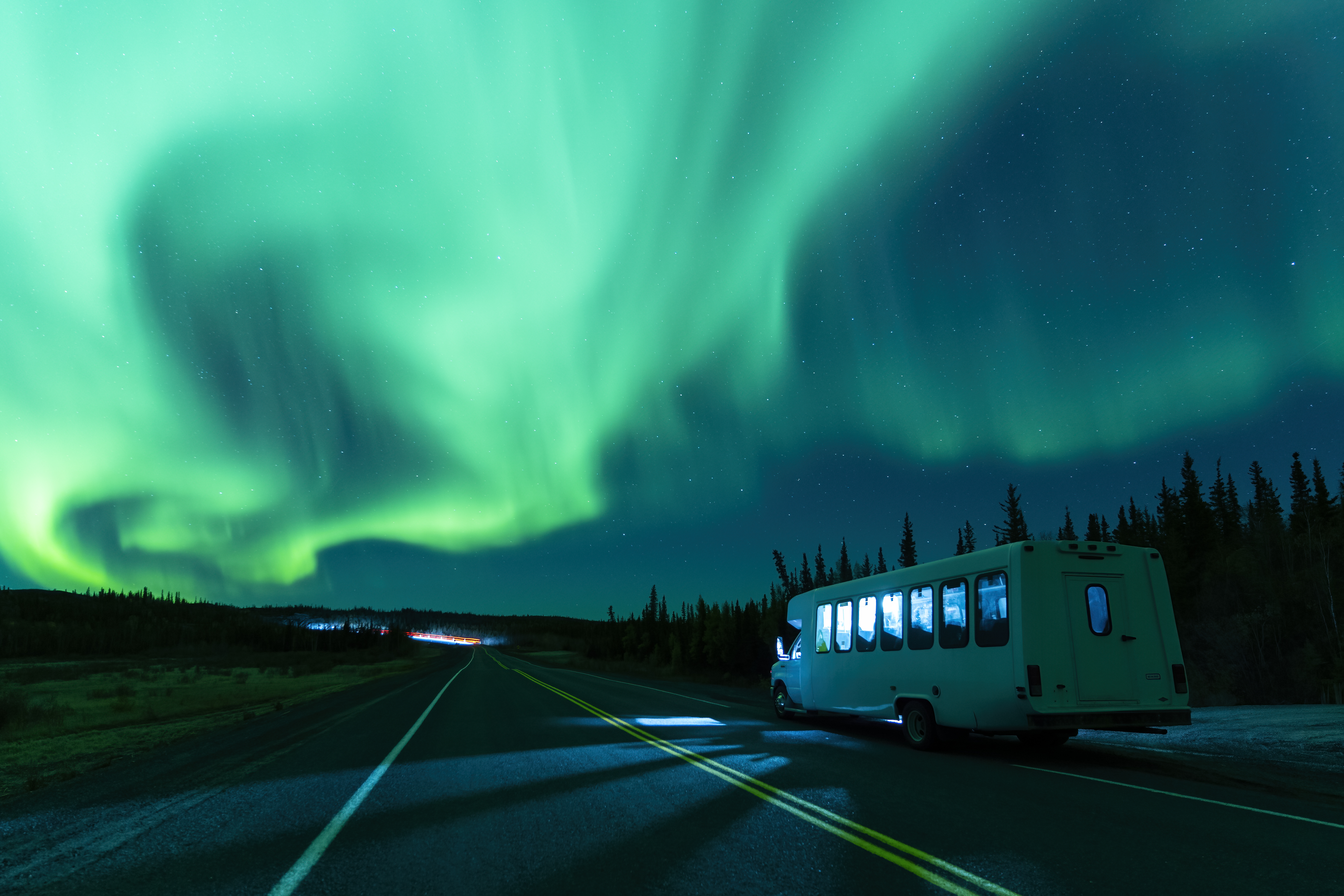 Small minibus drives along a quiet road with green aurora lighting up the sky above Yellowknife