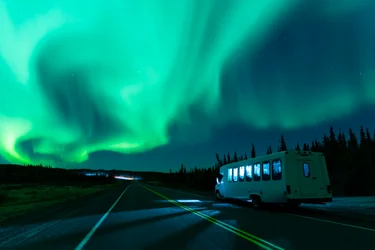 Small minibus drives along a quiet road with green aurora lighting up the sky above Yellowknife