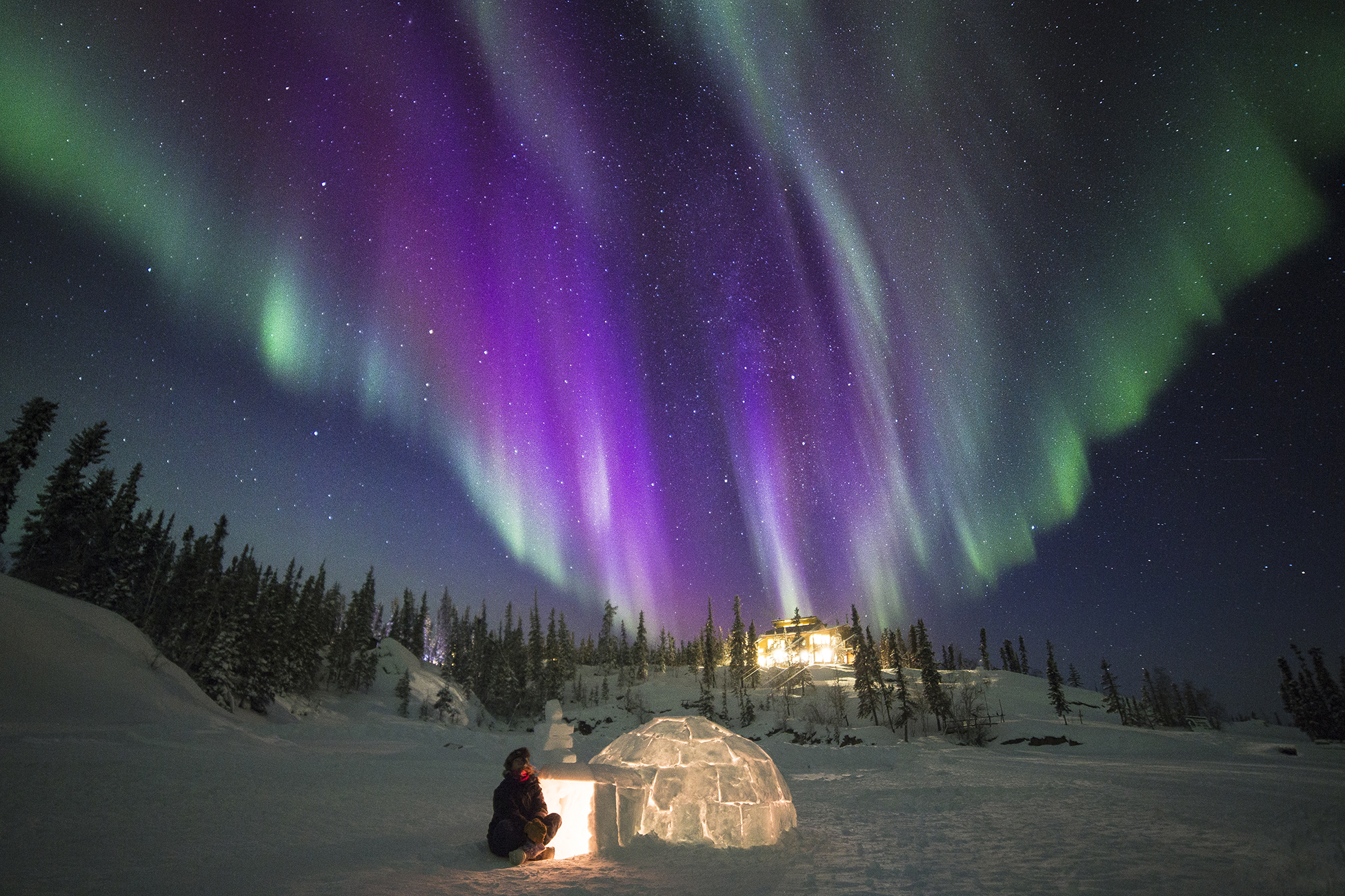 A person is sitting in front of an igloo under the aurora borealis