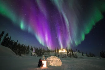 A person is sitting in front of an igloo under the aurora borealis