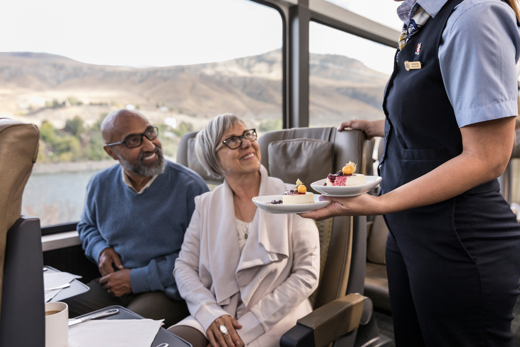 Seated senior couple smile at Rocky Mountaineer SilverLeaf train host as they are served dessert