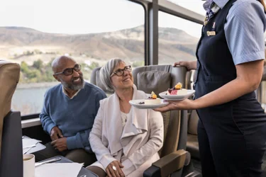 Seated senior couple smile at Rocky Mountaineer SilverLeaf train host as they are served dessert