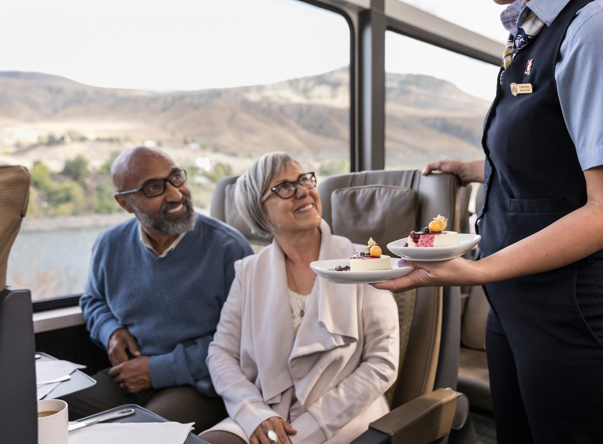 Seated senior couple smile joyfully as host faces the pair while serving dessert onboard Rocky Mountaineer SilverLeaf coach