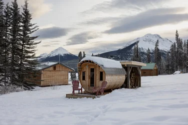 Barrel sauna in front of log cabins at the Southern Lakes Resort, snow on the ground and mountains behind
