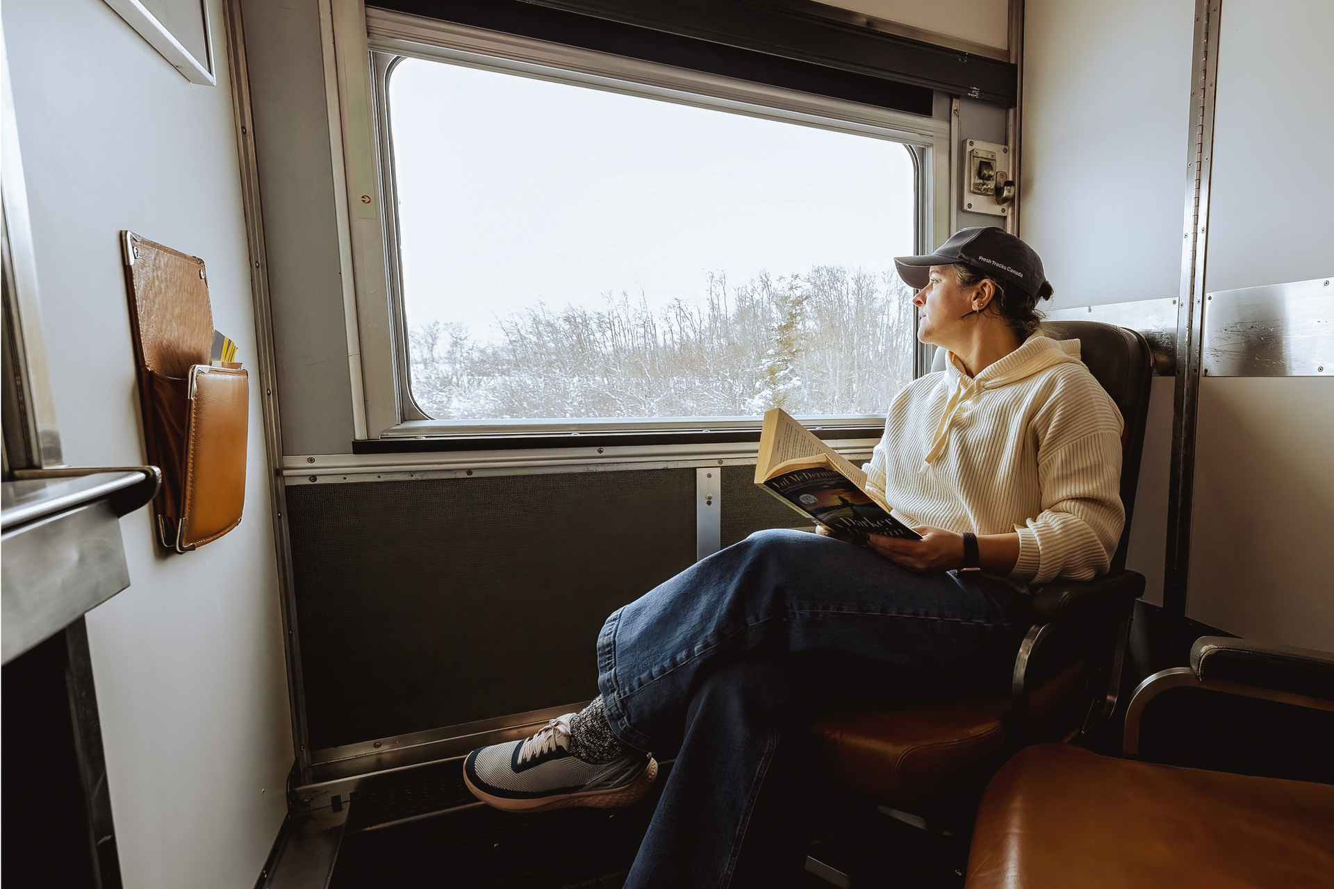 Mature woman seated in chair relaxes with a book in Sleeper Plus class cabin for two and looks at landscape from large window onboard VIA Rail train, the Canadian