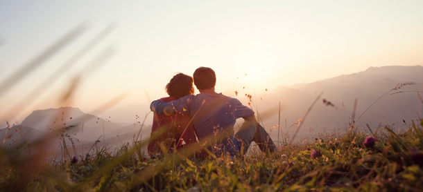 A couple sitting on a grassy slope stares out at the sunset