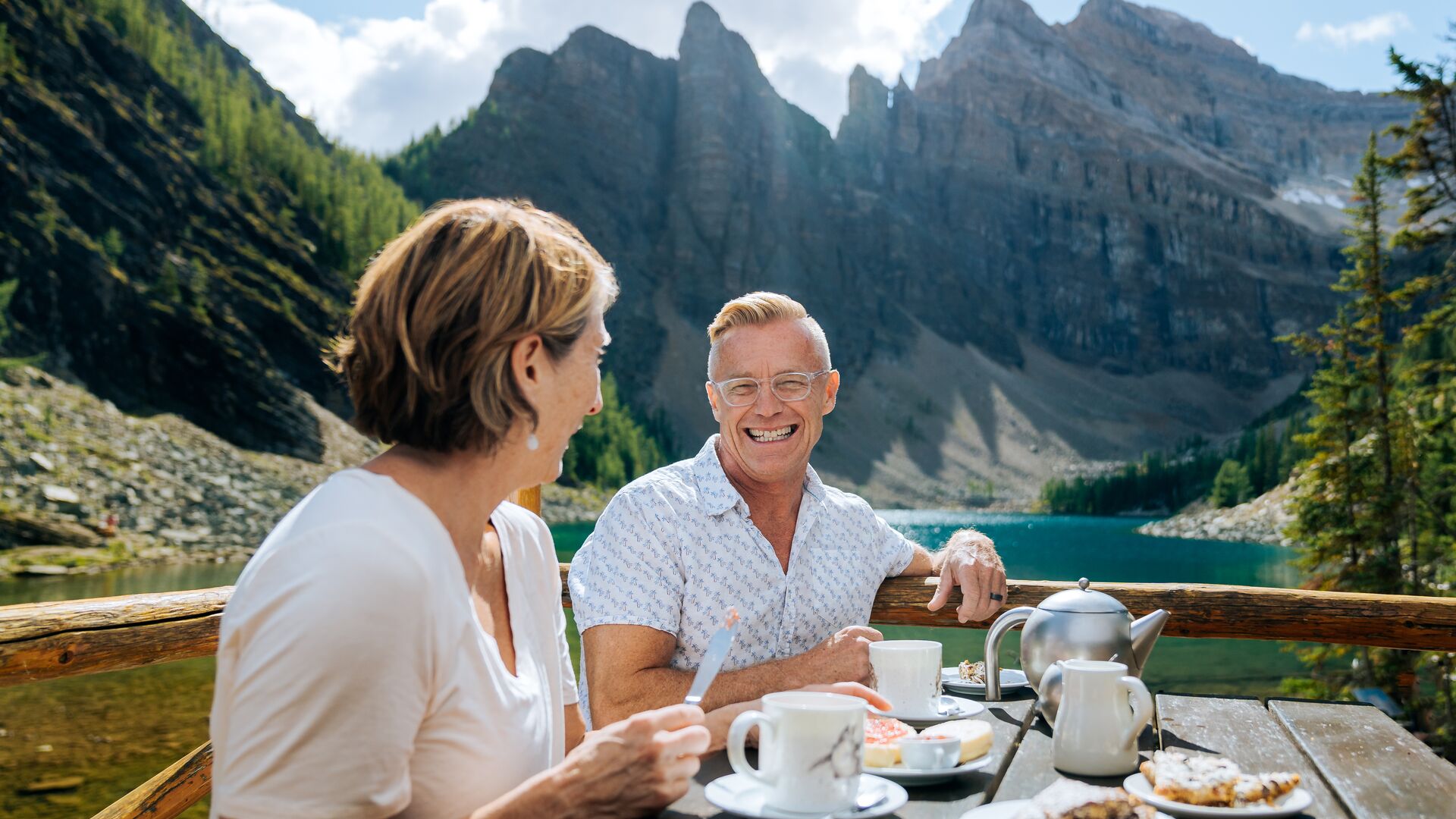 A couple having hot tea and baked goods on a patio with mountains in the background at Lake Agnes Tea House in Lake Louise, Alberta, Canada