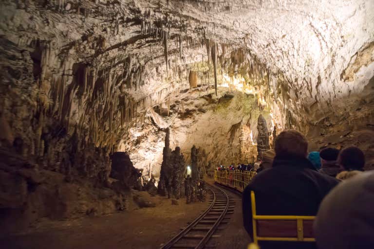 Illuminated stalactites hang from ceiling as tourists ride underground train through Postojna Cave's dramatic limestone formations