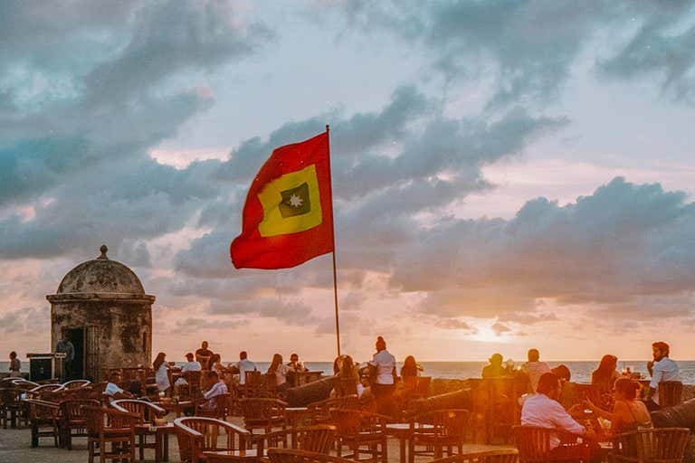 People gather at Café del Mar in Cartagena during sunset with a flag waving.
